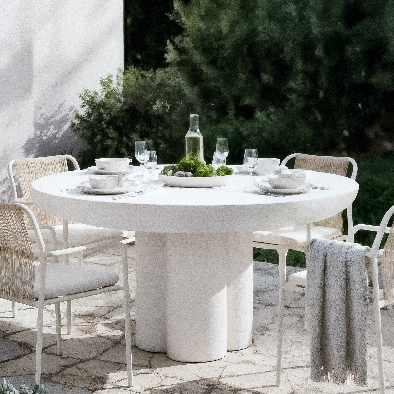 The Oberon 120cm Round Outdoor Concrete Dining Table in White is elegantly set with dinnerware and surrounded by four woven-seat chairs, on a stone-paved patio with lush greenery backdrop.