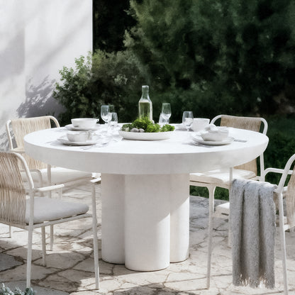The Oberon 120cm Round Outdoor Concrete Dining Table in White is elegantly set with dinnerware and surrounded by four woven-seat chairs, on a stone-paved patio with lush greenery backdrop.