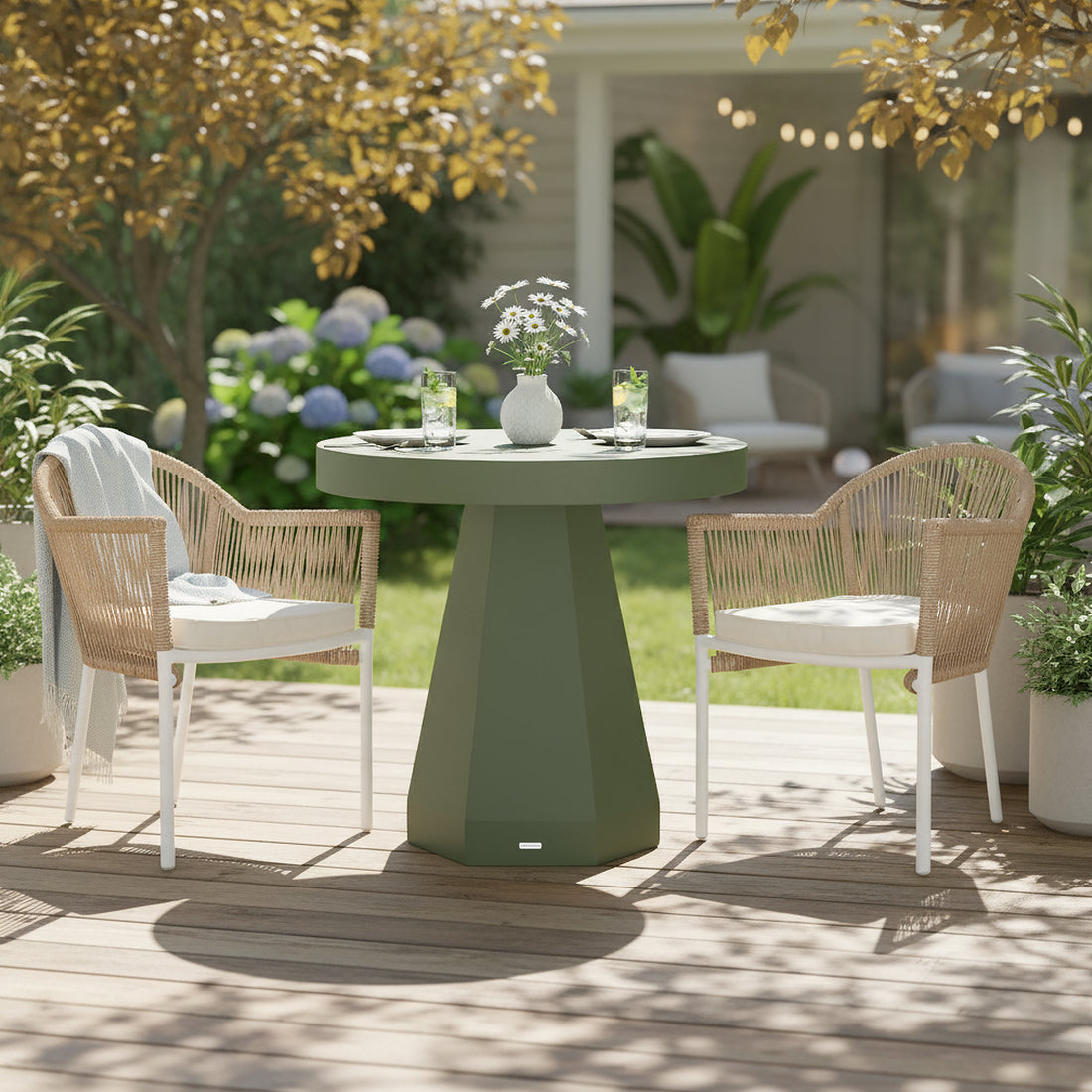 Orson 80cm Round Concrete Dining Table in Green on a wooden deck, topped with a white vase of daisies and two glasses, flanked by two woven-rope armchairs with cushions.