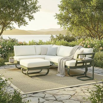 Paloma Modular Outdoor Lounge Set in Olive Green on a stone terrace, featuring an L-shaped sectional with pale cream cushions, a matching ottoman, and a small side table with a mug and book.