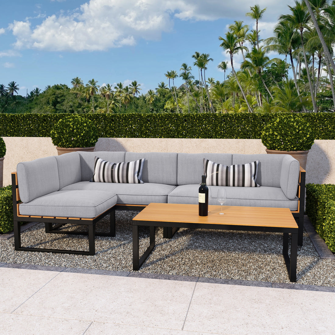 Panaluu 5 Seater Aluminium Corner Outdoor Lounge in Grey, featuring an L-shaped sofa with light-gray cushions, striped pillows, a matching coffee table, and a backdrop of lush greenery and palm trees.
