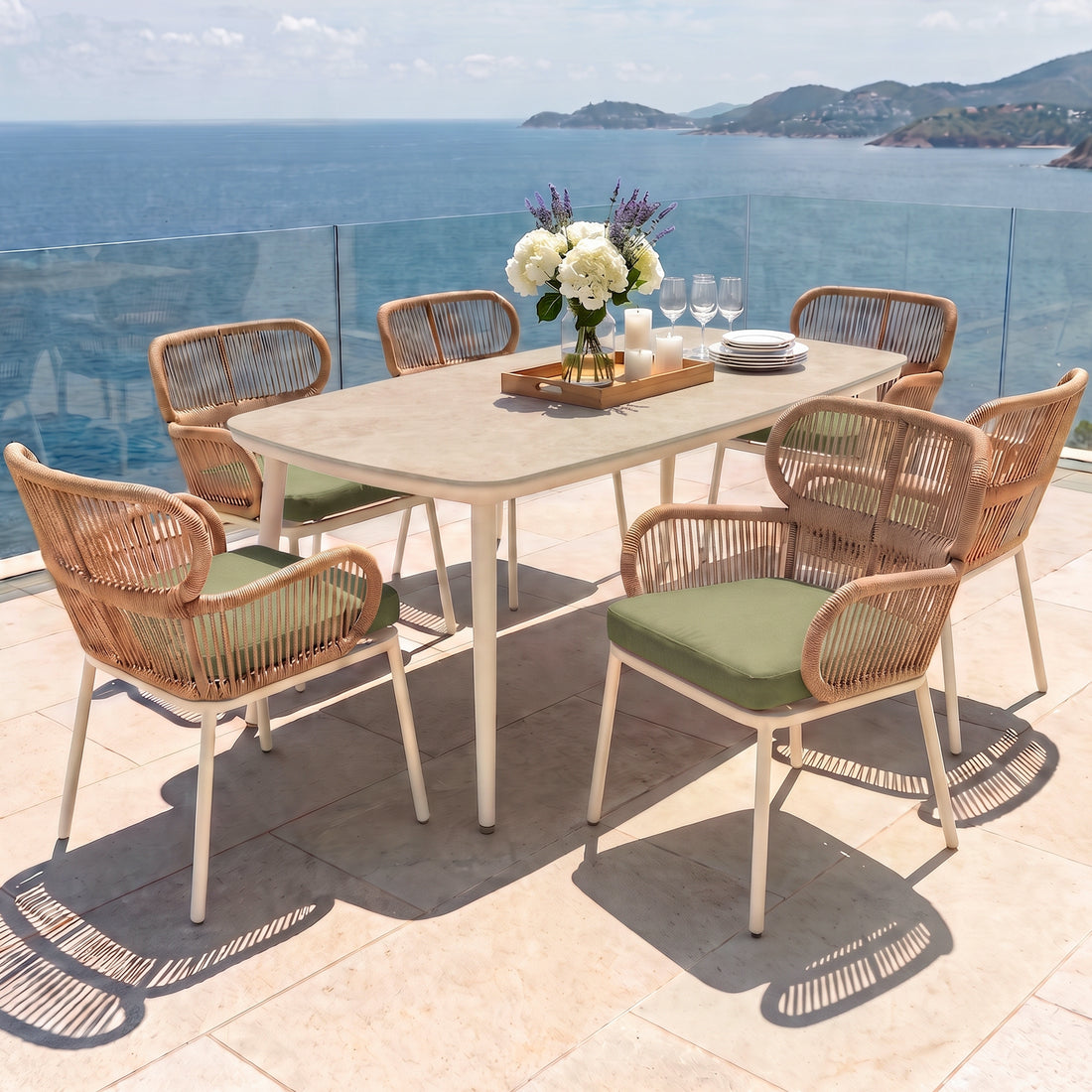 An outdoor dining setting on a terrace overlooking the sea, featuring a light-colored table with a centerpiece tray, candles, and flowers, surrounded by woven chairs with green cushions.