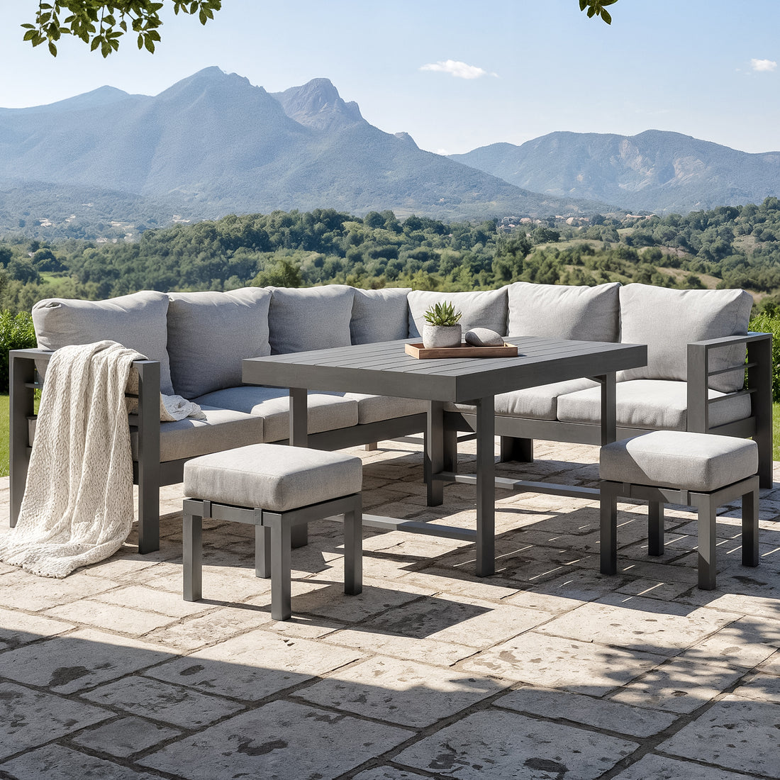 Portland Outdoor Corner Sofa Lounge with Footstools in Grey, featuring a modular L-shaped design with light gray cushions, a matching coffee table, and cushioned stools on a stone patio overlooking scenic hills.