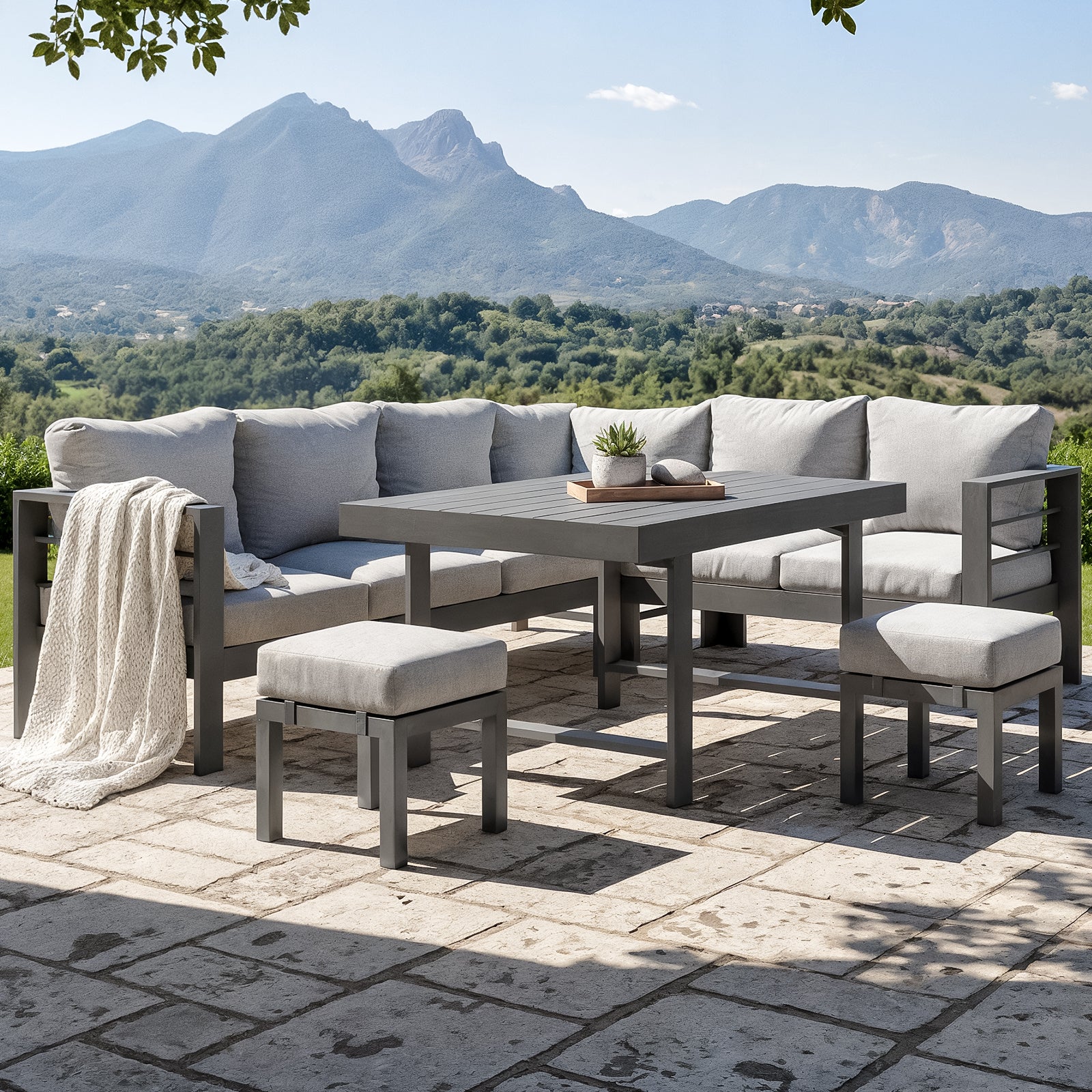 Portland Outdoor Corner Sofa Lounge with Footstools in Grey, featuring a modular L-shaped design with light gray cushions, a matching coffee table, and cushioned stools on a stone patio overlooking scenic hills.