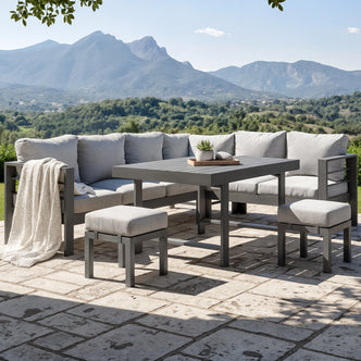 Portland Outdoor Corner Sofa Lounge with Footstools in Grey, featuring a modular L-shaped design with light gray cushions, a matching coffee table, and cushioned stools on a stone patio overlooking scenic hills.