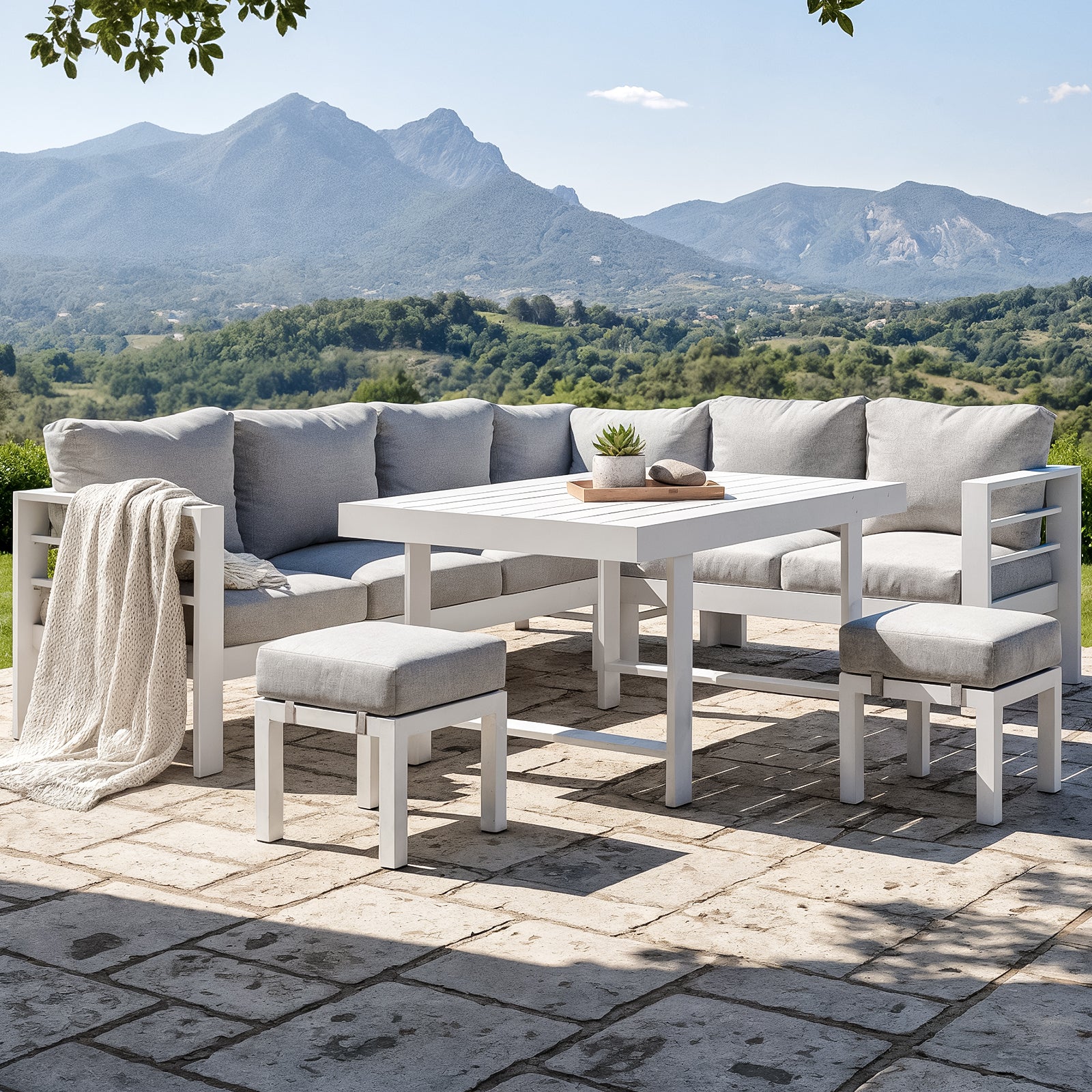 Portland Outdoor Corner Sofa Lounge with Footstools in White & Grey, featuring an L-shaped sectional, two ottomans, and a table with a succulent, set against scenic rolling hills and mountains.