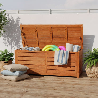 Luxo 150cm Eucalyptus Wood Outdoor Storage Box in Natural with open lid, showing towels and inflatable rings inside. Surrounded by potted plants and outdoor cushions, set on a wood-plank deck.