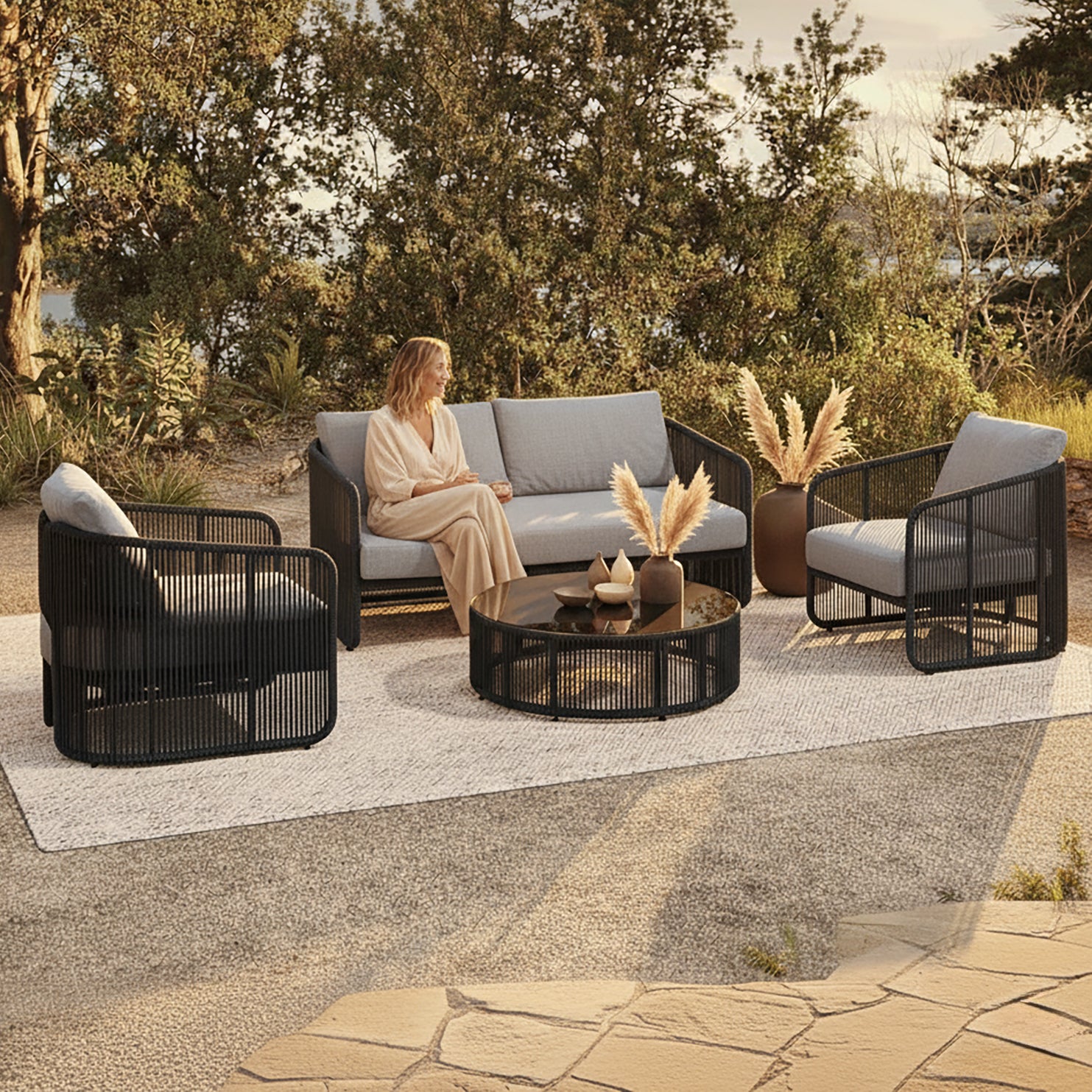 Solari 4 Seater Wicker Outdoor Sofa Set in Black features a round coffee table, loveseat, and two armchairs with plush gray cushions, set in a sunlit garden.
