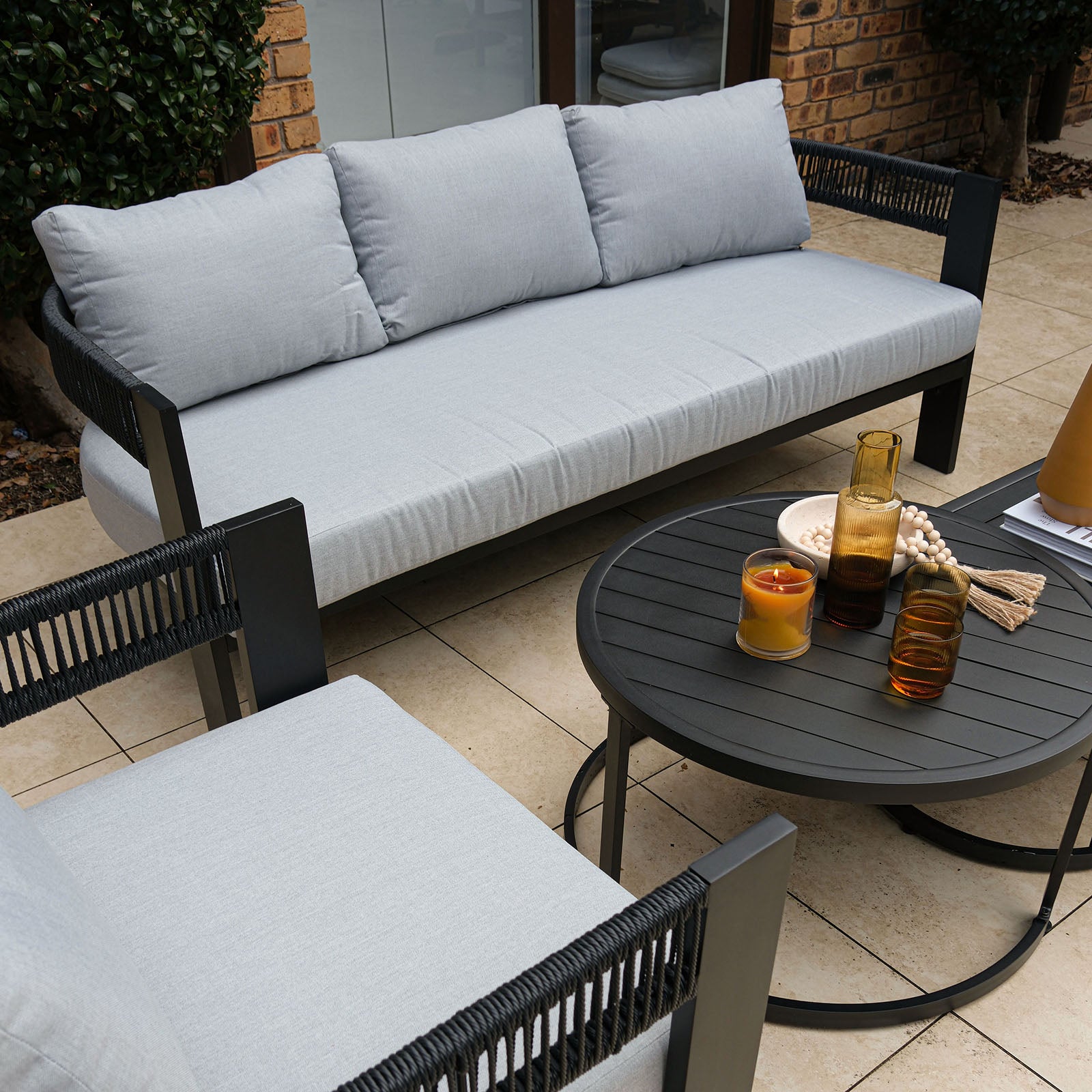 Sorrelle 5 Seater Wicker Outdoor Sofa Set in Black on a patio, featuring light gray cushions, a round coffee table with amber glassware, and a decorative bowl, set against a brick wall.