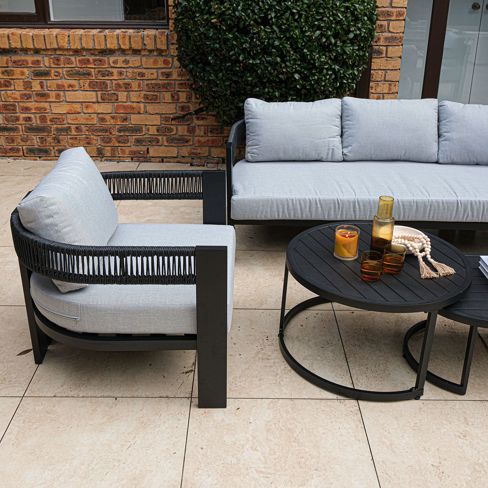 Sorrelle 5 Seater Wicker Outdoor Sofa Set in Black on a stone patio, featuring a black metal armchair and three-seat sofa with gray cushions, paired with black nesting coffee tables holding decor.