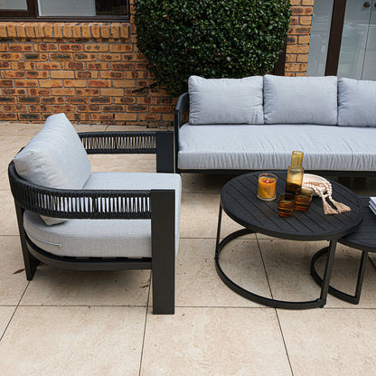 Sorrelle 5 Seater Wicker Outdoor Sofa Set in Black on a stone patio, featuring a black metal armchair and three-seat sofa with gray cushions, paired with black nesting coffee tables holding decor.