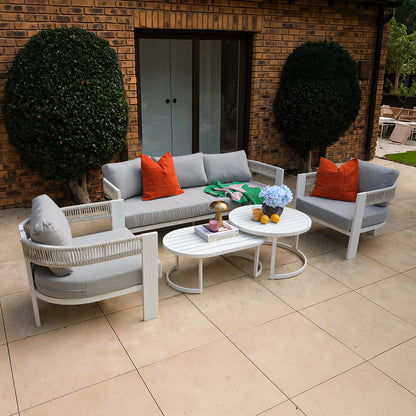 Sorrelle 5 Seater Wicker Outdoor Sofa Set in Light Grey with orange pillows, white rope accents, two oval tables, blue hydrangeas, books, and a gold lamp on stone pavers.