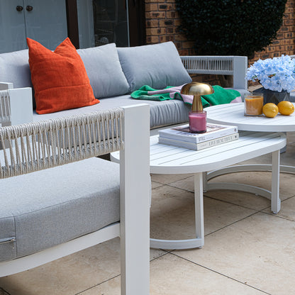 Sorrelle 5 Seater Wicker Outdoor Sofa Set in Light Grey featuring a white-framed sofa with cushions, burnt orange pillow, green/pink blanket, and two nested coffee tables with décor items.