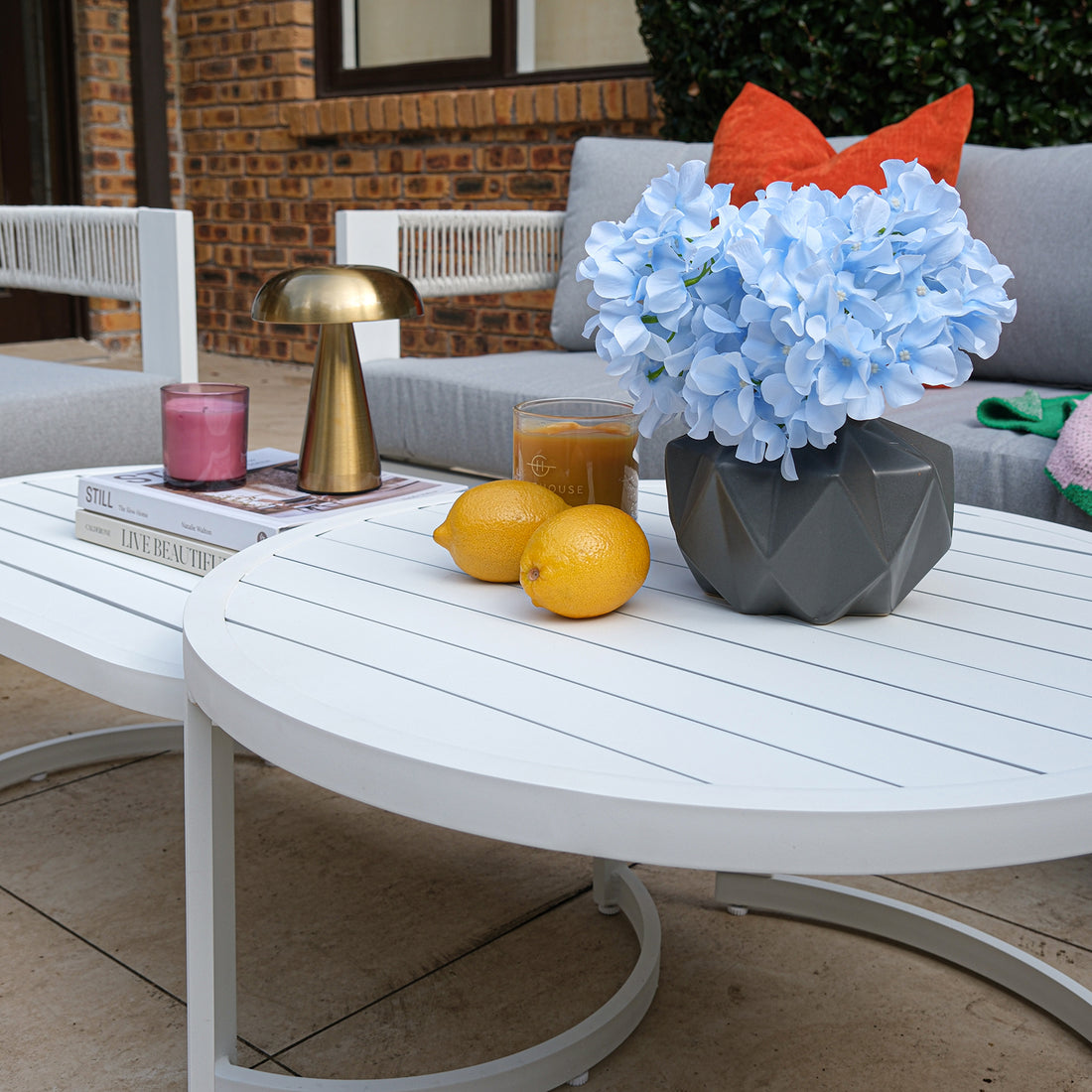 Sorrelle 5 Seater Wicker Outdoor Sofa Set in Light Grey features a round white coffee table with hydrangeas, lemons, iced coffee, a candle, and books in a stylish outdoor setting.
