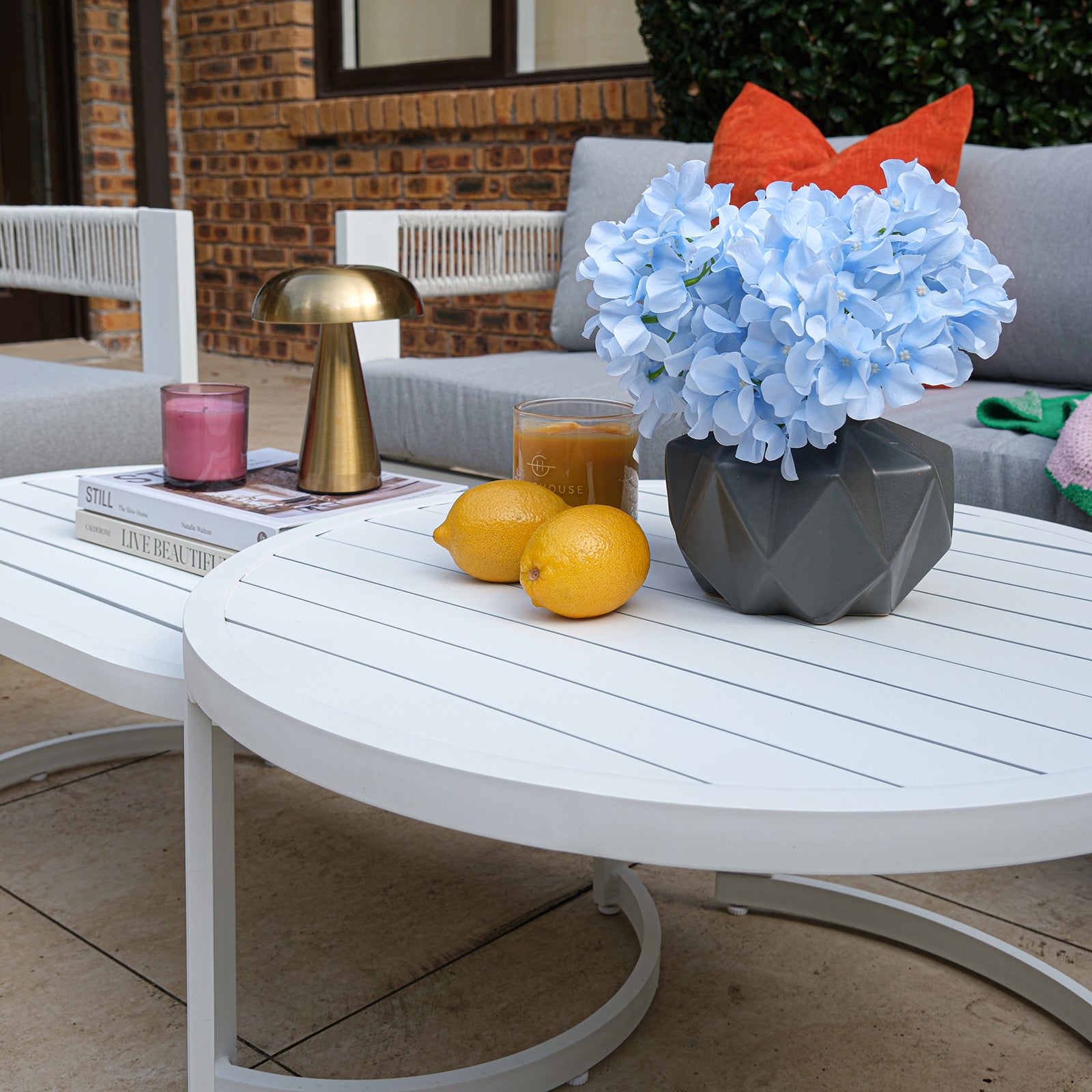 Sorrelle 5 Seater Wicker Outdoor Sofa Set in Light Grey features a round white coffee table with hydrangeas, lemons, iced coffee, a candle, and books in a stylish outdoor setting.