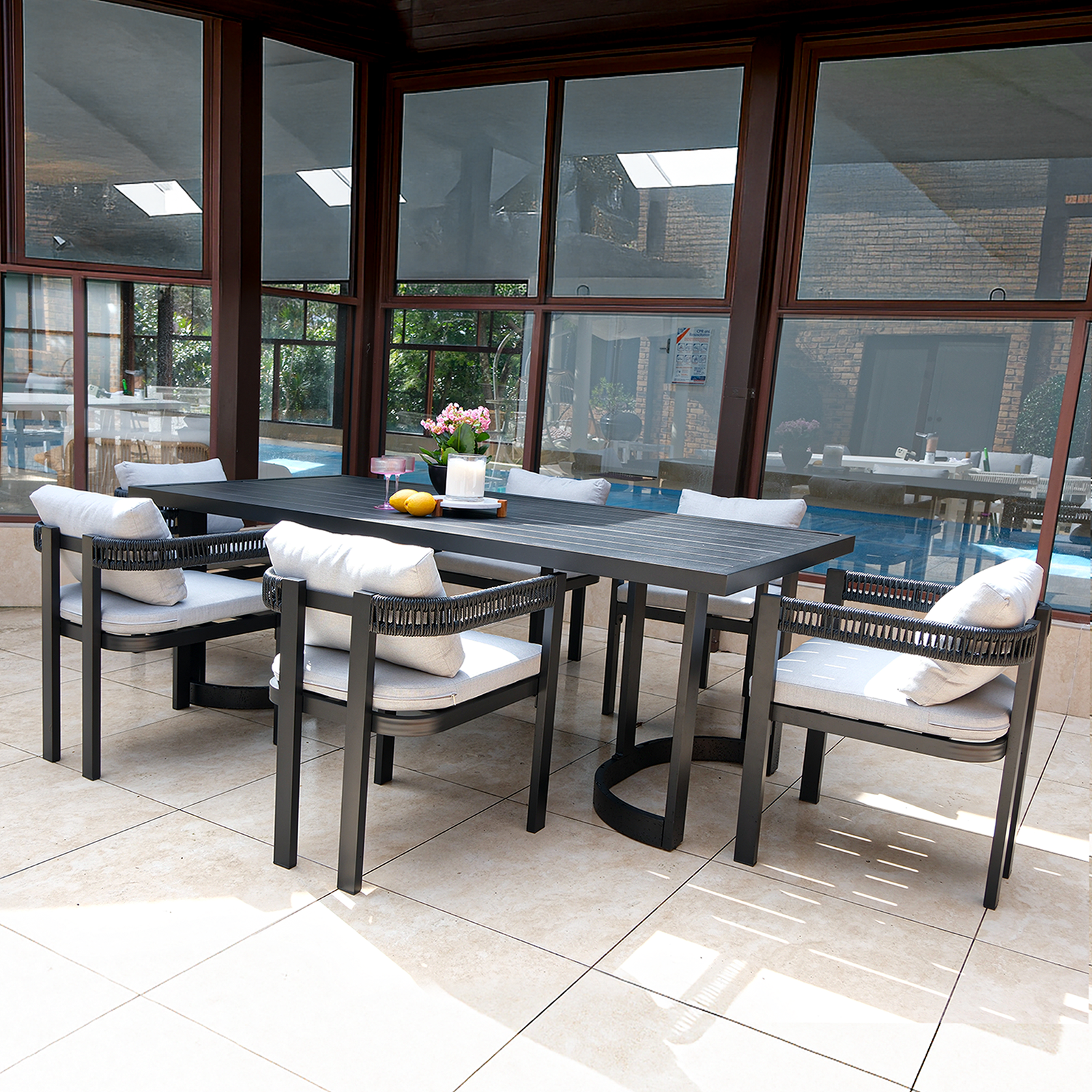 Tavari 6-Seater Aluminium Outdoor Dining Set in Black features a sleek black table and six chairs with light-gray cushions, set in a modern patio by a pool, accented with decor elements.