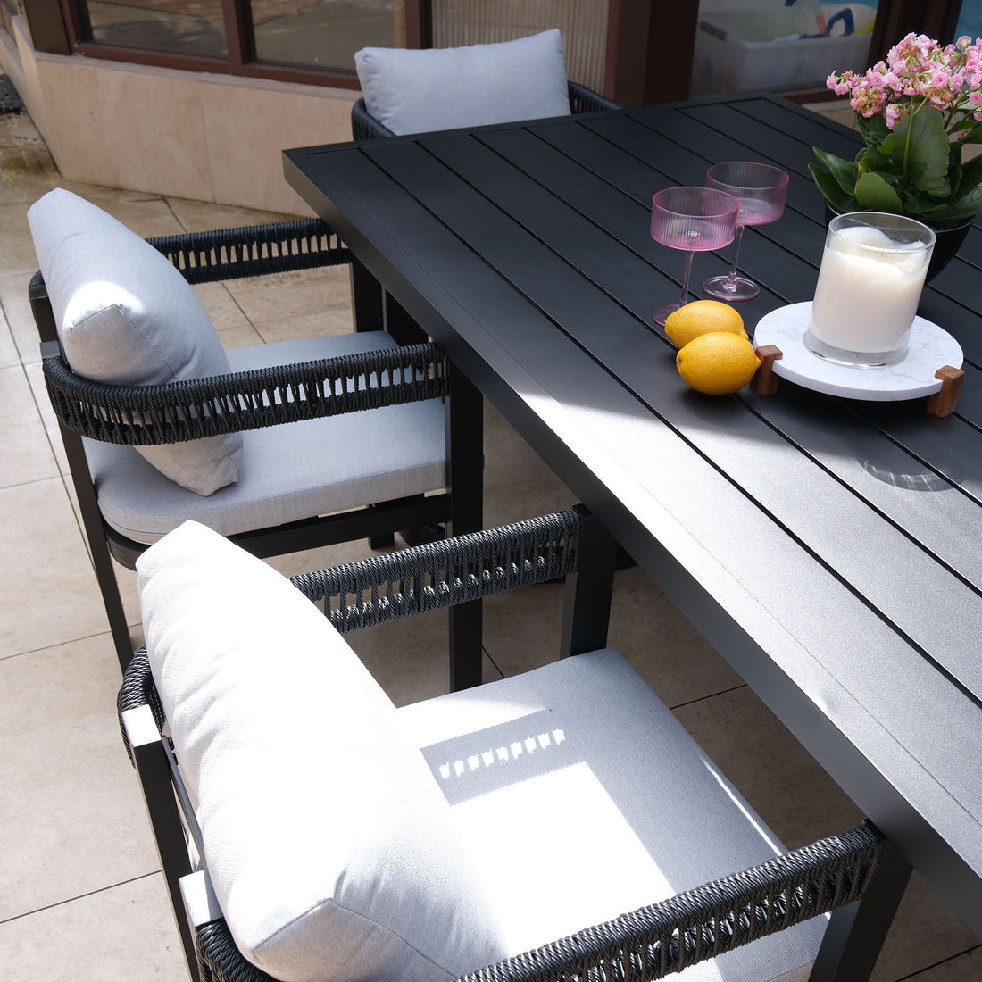 Tavari 6-Seater Aluminium Outdoor Dining Set in Black featuring a black metal-slatted table, two woven-rope armchairs with cushions, a potted plant, candle, lemons, and pink glasses on a tiled patio.