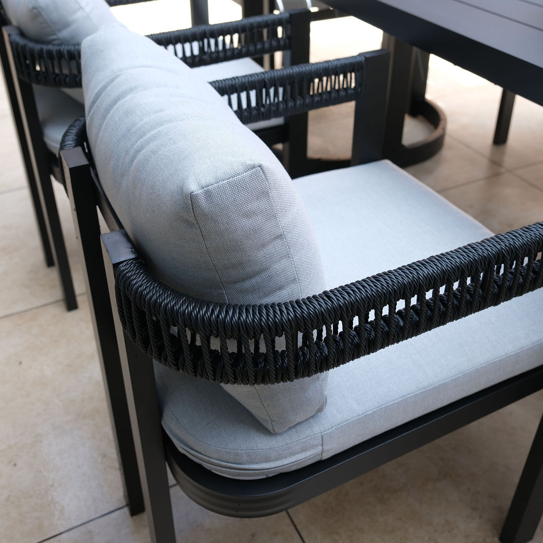 Tavari 6-Seater Aluminium Outdoor Dining Set in Black, featuring a close-up of a chair with a black metal frame, woven rope armrests, and light-grey cushions on a tiled patio.