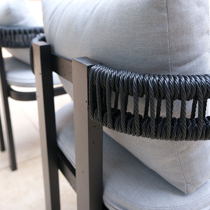 Tavari 6-Seater Aluminium Outdoor Dining Set in Black features a close-up of a black metal-framed chair with black rope backrest, light gray cushions; a second chair is partially visible behind.