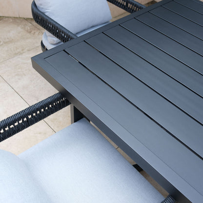 Close-up of Tavari 6-Seater Aluminium Outdoor Dining Set in Black, showing a dark slatted metal table corner and matching chair with woven rope arms and light-gray cushions on a tiled patio.