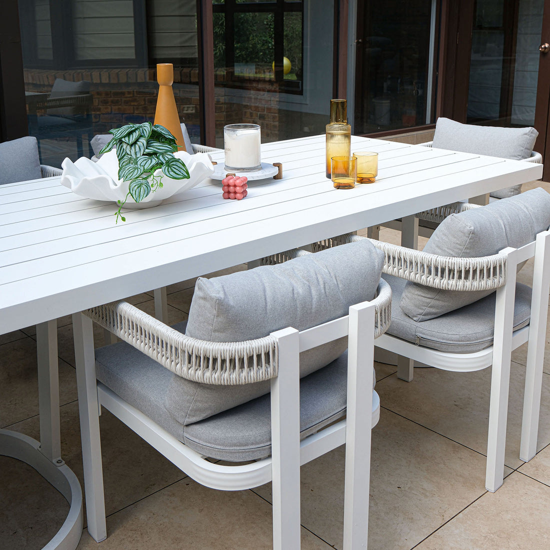 Tavari 6-Seater Aluminium Outdoor Dining Set in Light Grey featuring a white slatted table, four white-framed chairs with woven rope backs, and decorative items on a light beige tiled patio.