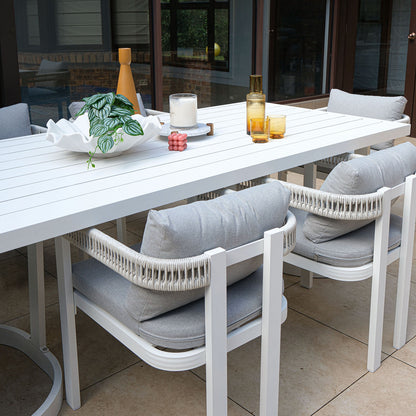 Tavari 6-Seater Aluminium Outdoor Dining Set in Light Grey featuring a white slatted table, four white-framed chairs with woven rope backs, and decorative items on a light beige tiled patio.