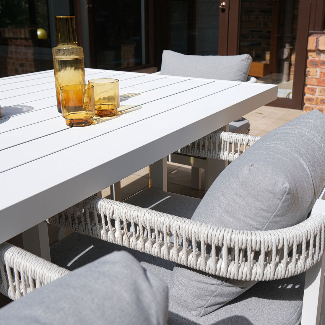 Tavari 6-Seater Aluminium Outdoor Dining Set in Light Grey features a white slatted table with amber carafe and tumblers, surrounded by woven-rope chairs with light grey cushions, perfect for stylish outdoor gatherings.