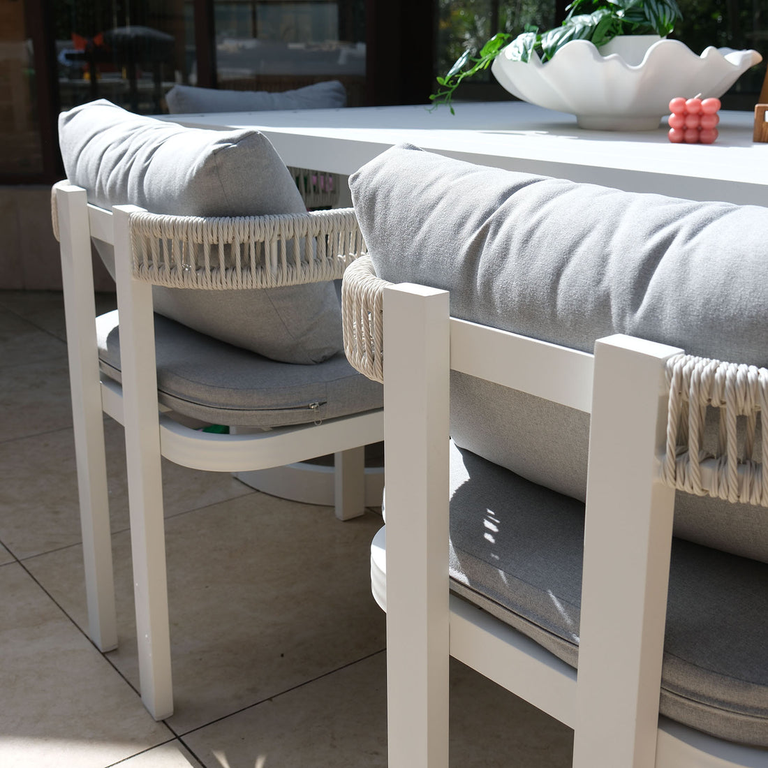 Tavari 6-Seater Aluminium Outdoor Dining Set in Light Grey featuring two white woven rope-style chairs with cushions, a white tabletop, a scalloped bowl with a plant, and a pink decorative object.