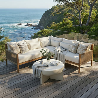 Tavi 5 Seater Solid Timber L Shape Outdoor Sofa in Beige features a teak-like frame with off-white cushions, accented by various throw pillows and a knit throw, set against a coastal backdrop.