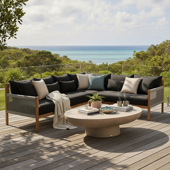 Tavi 5 Seater Solid Timber L Shape Outdoor Sofa with woven rope sides, deep cushions, and throw pillows on a seaside deck, accompanied by a stone-look coffee table with decor items.