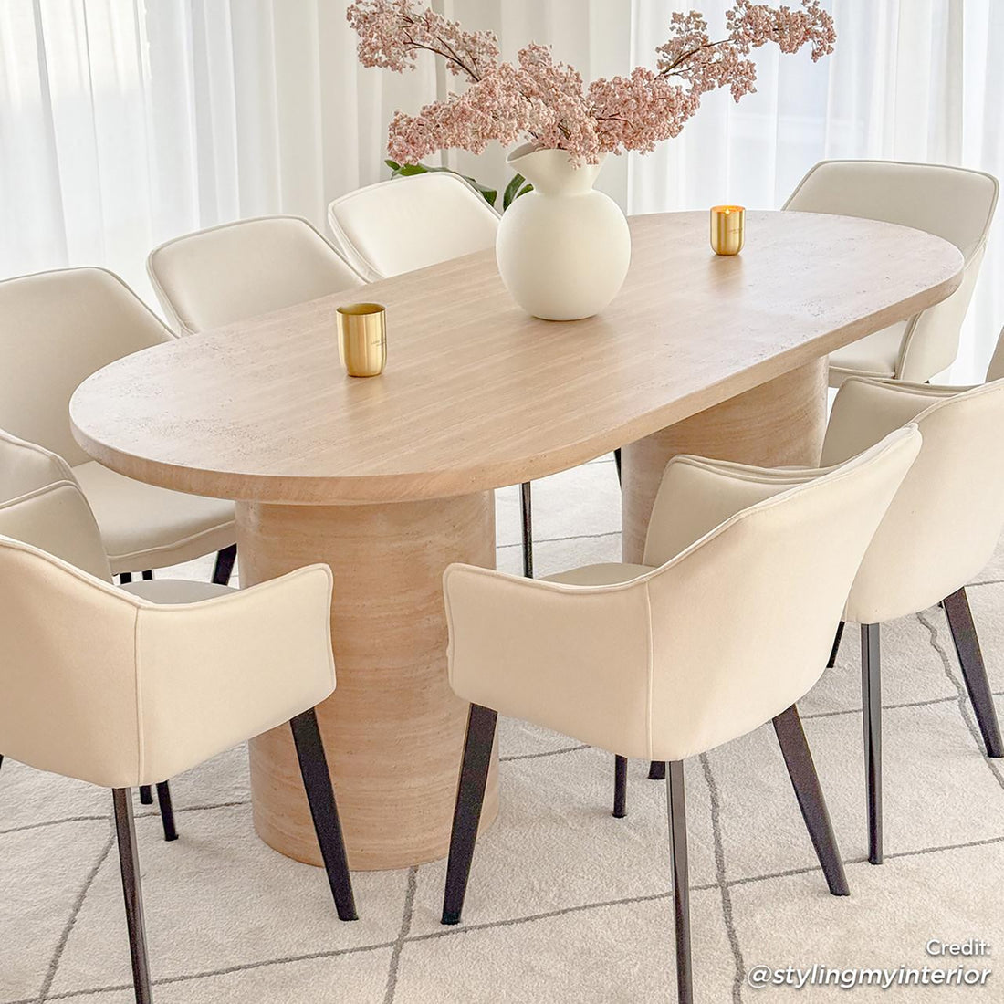 Tierre 200cm Concrete Indoor/Outdoor Dining Table in Beige, featuring an oval, light-wood design with cylindrical pedestals, surrounded by cream upholstered chairs, accented by a white vase with pink blossoms.