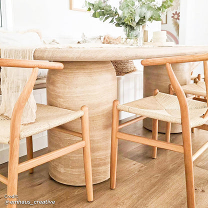 Tierre 200cm Round Outdoor Concrete Dining Table in Beige with a sand-colored pedestal base, topped with greenery in a clear vase, surrounded by oak-framed chairs with woven seats.