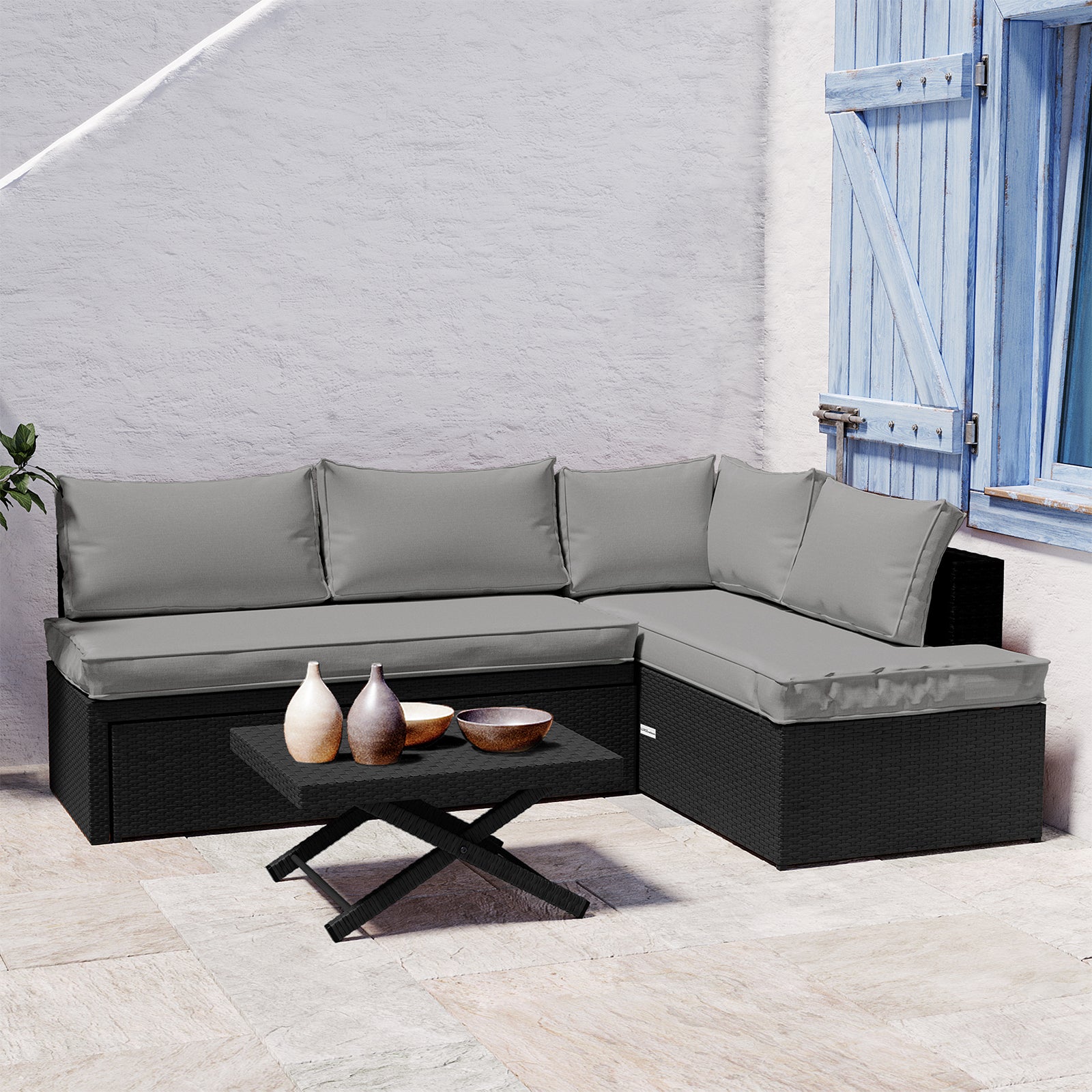 Tybee 4 Seater Rattan Outdoor Corner Sofa Set in Warm Grey features a black synthetic rattan sectional with light gray cushions and a matching coffee table, enhanced by decorative ceramic vases against a white stucco backdrop.