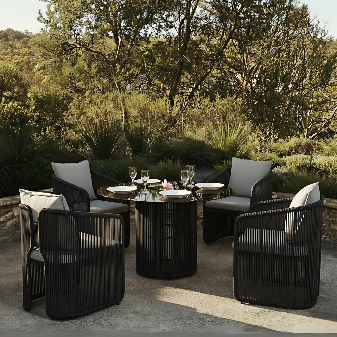 Vellora 4 Seater Wicker Outdoor Dining Set in Black features a round table with a glossy top and four dark woven armchairs, each with light gray cushions, set on a patio.