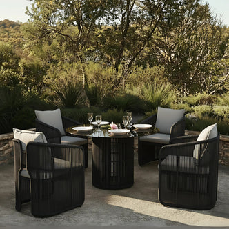 Vellora 4 Seater Wicker Outdoor Dining Set in Black features a round table with a glossy top and four dark woven armchairs, each with light gray cushions, set on a patio.