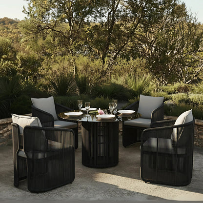 Vellora 4 Seater Wicker Outdoor Dining Set in Black features a round table with a glossy top and four dark woven armchairs, each with light gray cushions, set on a patio.