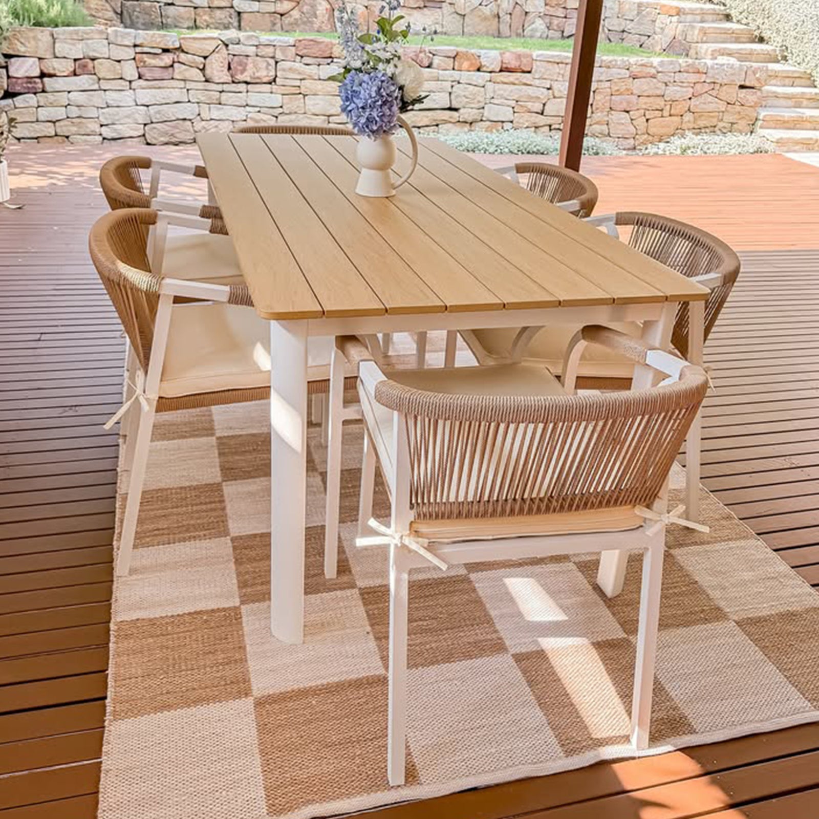 Vernon 6 Seater Wicker Outdoor Dining Set in Natural, featuring a light-wood table and six rope-backed chairs with seat pads on a checkered rug, under a covered wood-decked patio.