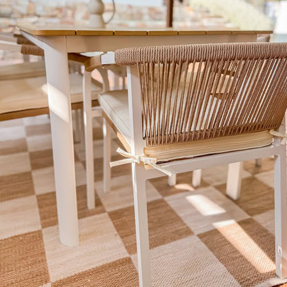 Vernon 6 Seater Wicker Outdoor Dining Set in Natural features a light wood table with white metal legs, surrounded by natural-tone rope-wrapped chairs with off-white cushions, set on a beige checkerboard rug.