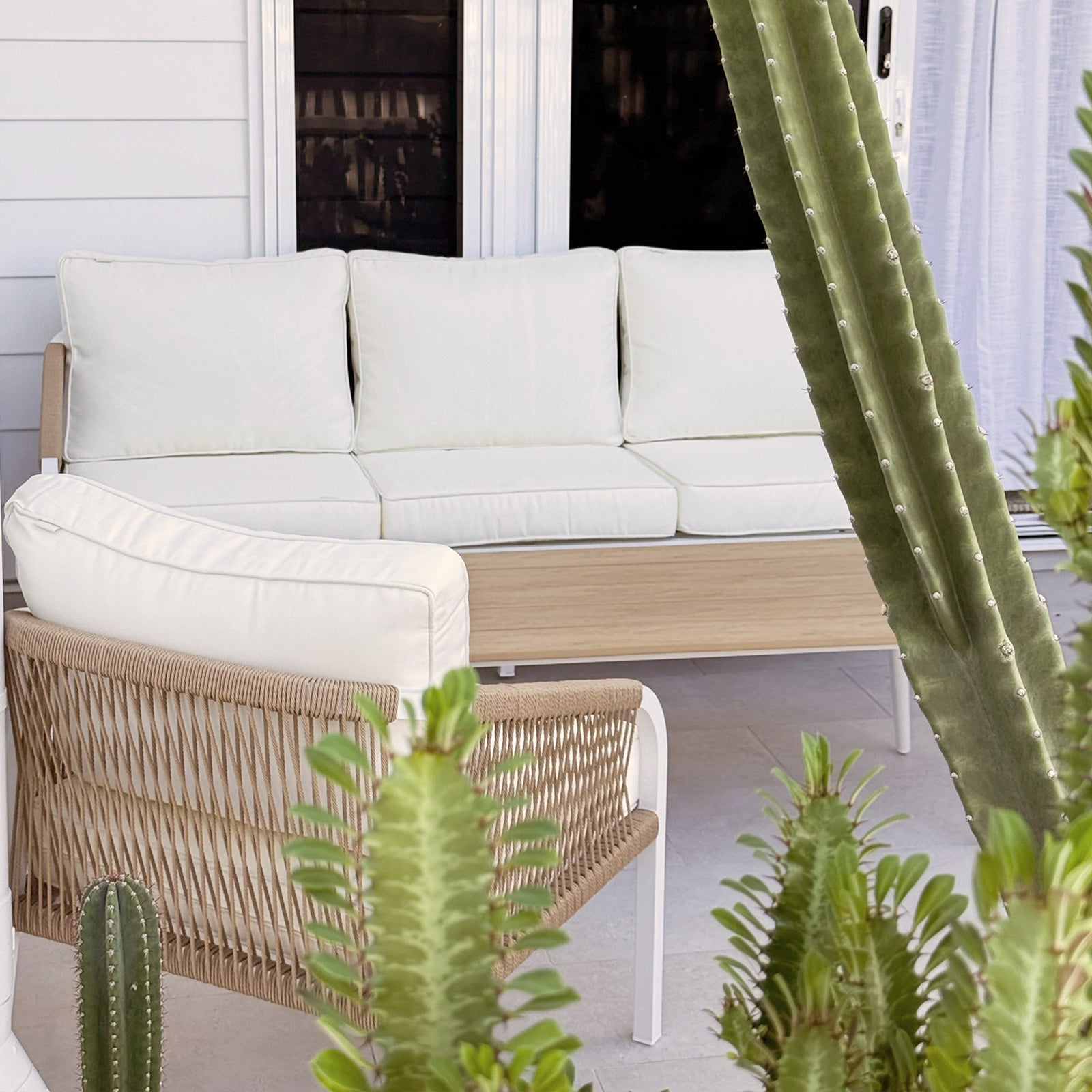 Vernon 5 Seater Aluminium Outdoor Sofa Set in Cream White, featuring a light-wood frame, white cushions, and woven rope armchair sides, nestled among tall succulents on a tiled patio.