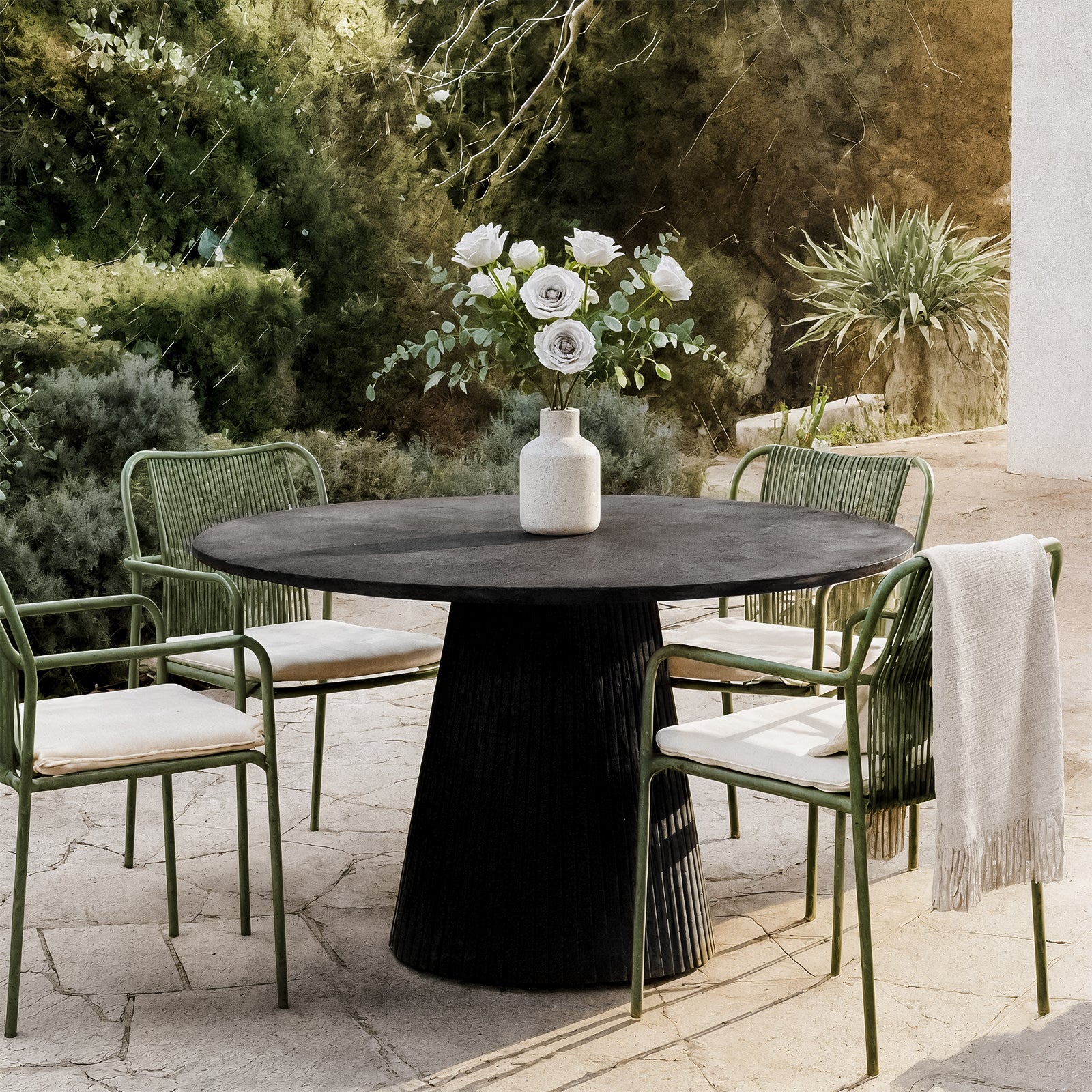 Xander 120cm Fluted Outdoor Concrete Dining Table Grey Marble, shown on a stone-tiled patio, features a vase with white roses and eucalyptus, surrounded by green metal-framed chairs with woven backs.