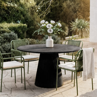 Xander 120cm Fluted Outdoor Concrete Dining Table Grey Marble, shown on a stone-tiled patio, features a vase with white roses and eucalyptus, surrounded by green metal-framed chairs with woven backs.