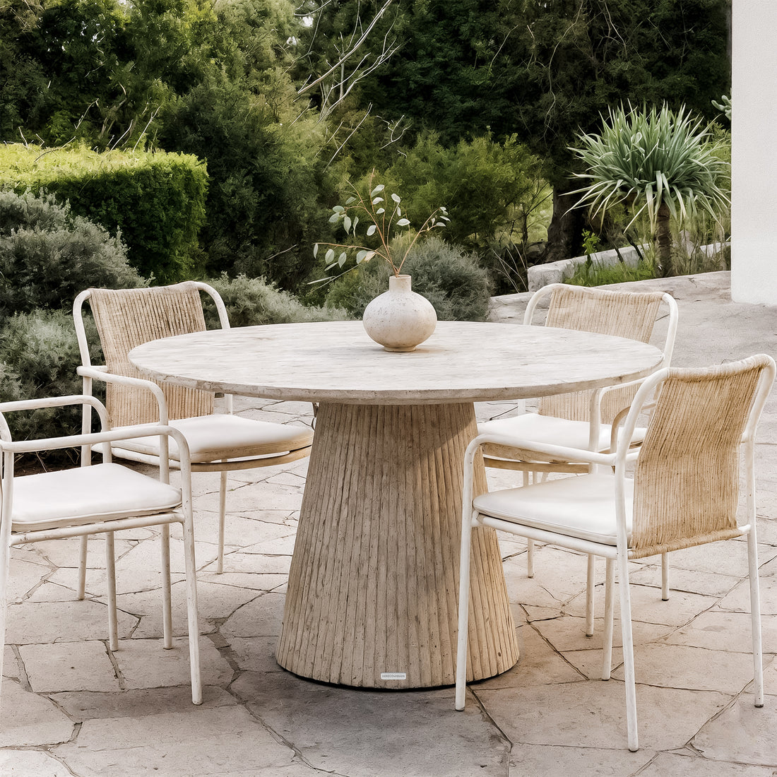 Xander 120cm Fluted Outdoor Concrete Dining Table in Ivory, featuring a round top and fluted pedestal, surrounded by four woven-back chairs on a stone-slab patio with lush greenery in the background.