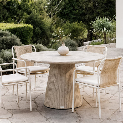 Xander 120cm Fluted Outdoor Concrete Dining Table in Ivory, featuring a round top and fluted pedestal, surrounded by four woven-back chairs on a stone-slab patio with lush greenery in the background.