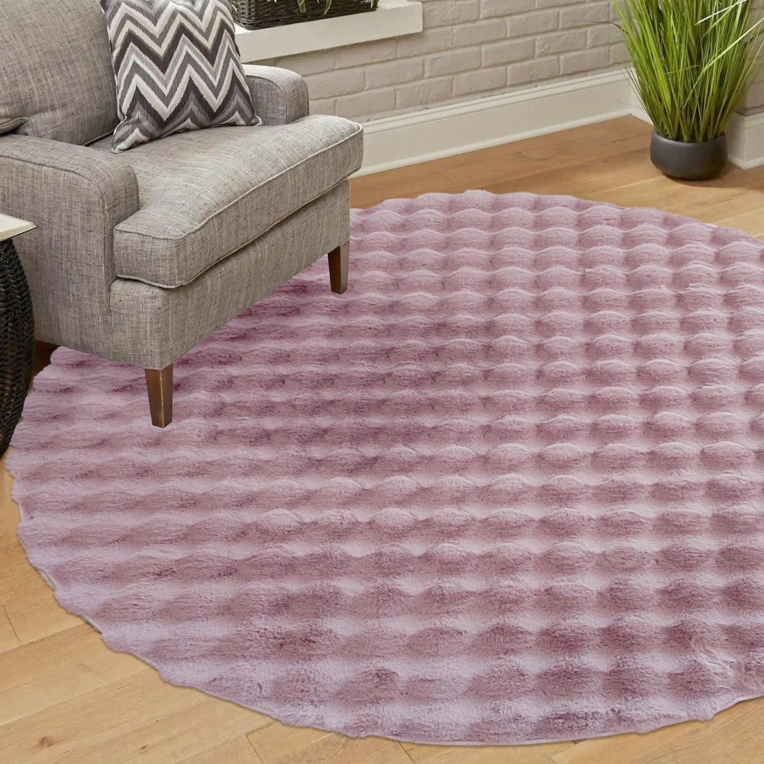 Armida 160cm Round Fluffy Faux Fur Bubble Rug in Dusty Pink, featuring a plush, shaggy texture, placed under a gray armchair with wooden legs and a chevron-patterned pillow.