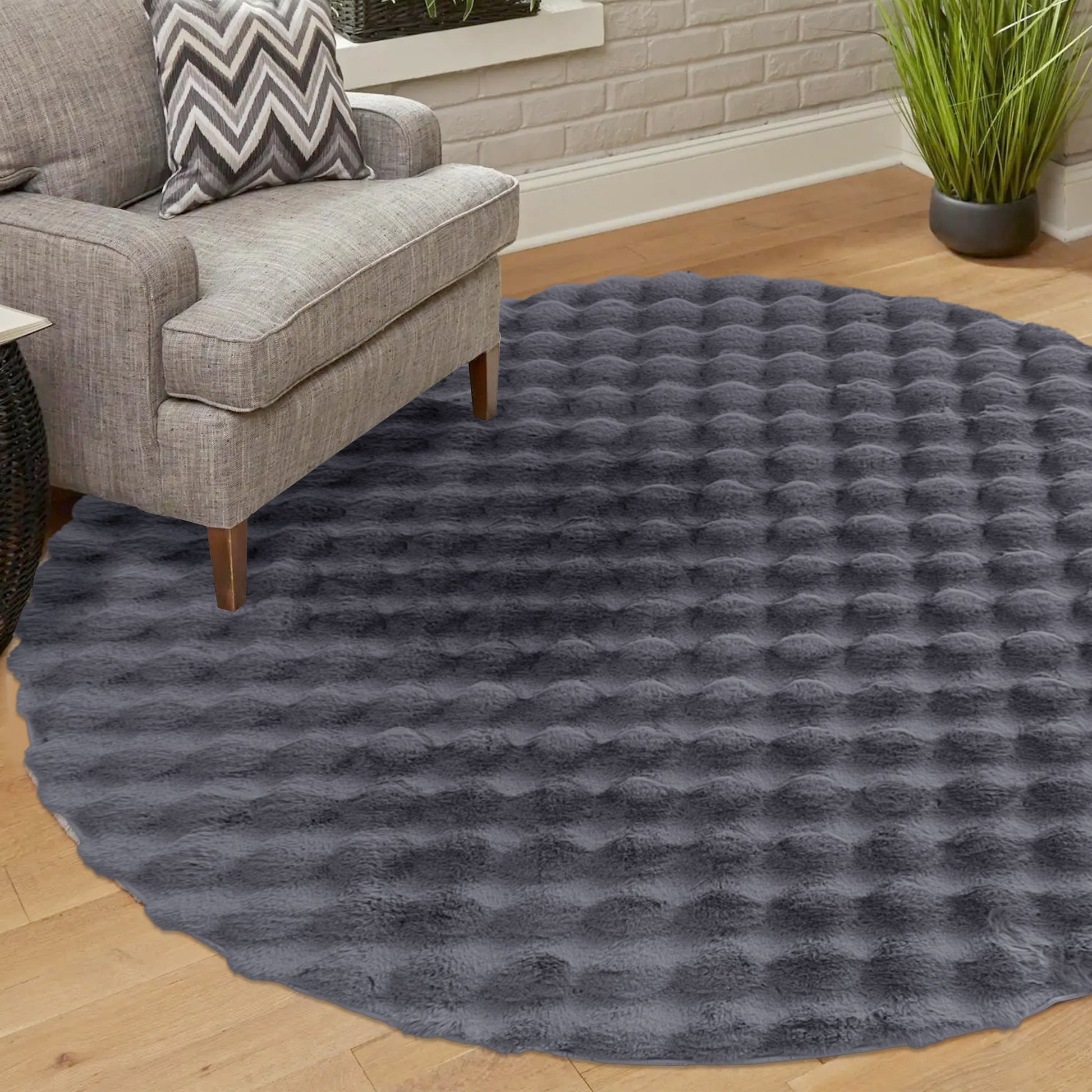 Armida 160cm Round Fluffy Faux Fur Bubble Rug in Grey with a shaggy bubble texture, partially covered by a light gray upholstered armchair and chevron-patterned pillow, set on a wooden floor.