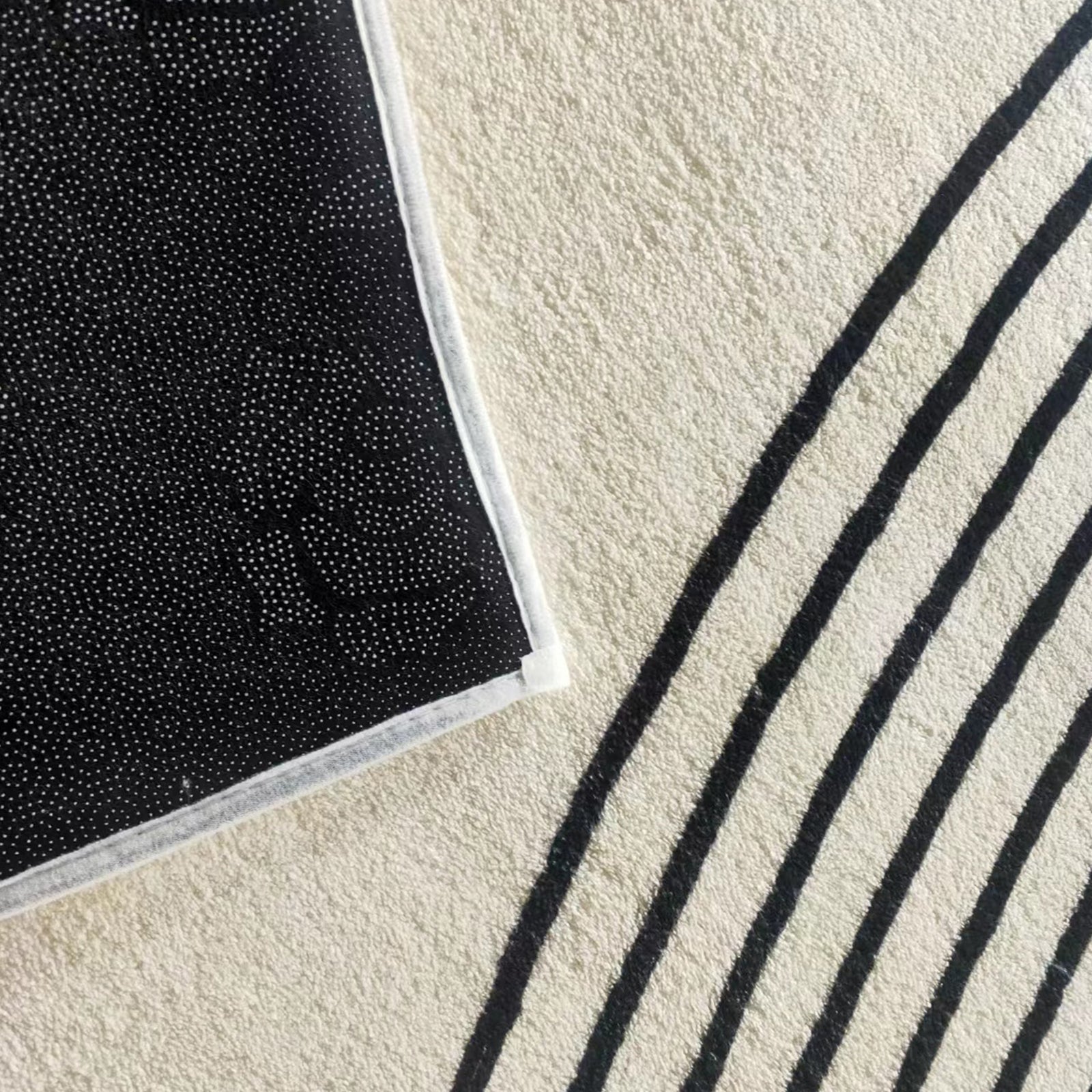Close-up of Axland Modern Abstract Rug 120x180cm, showing light-beige surface with thin, curved black stripes, slightly overlapping with black, grip-dotted underside of another rug or rug pad.