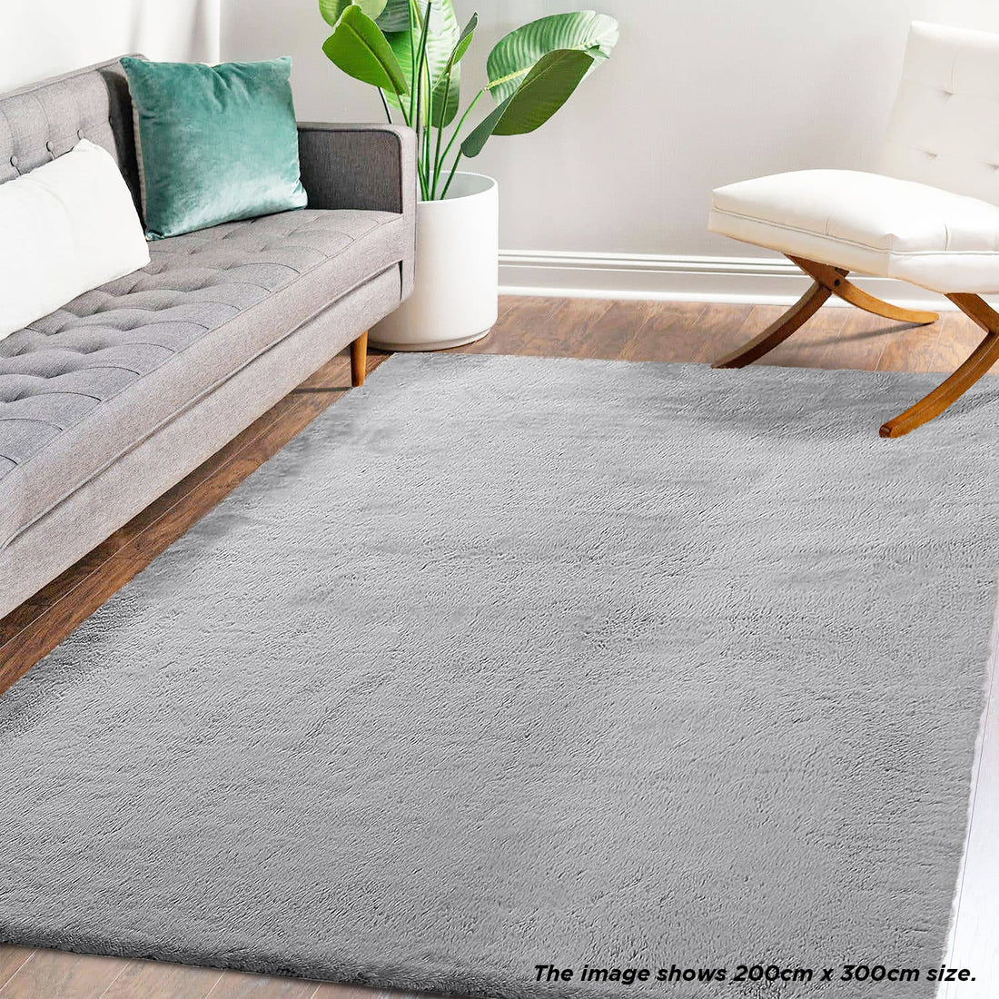 Azaliya 200x300cm Faux Rabbit Fur Floor Rug in silver, covering a wood-tone floor in a modern living area with a gray sofa, green pillow, white chair, and houseplant.