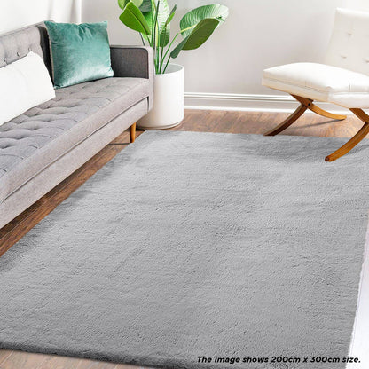 Azaliya 200x300cm Faux Rabbit Fur Floor Rug in silver, covering a wood-tone floor in a modern living area with a gray sofa, green pillow, white chair, and houseplant.