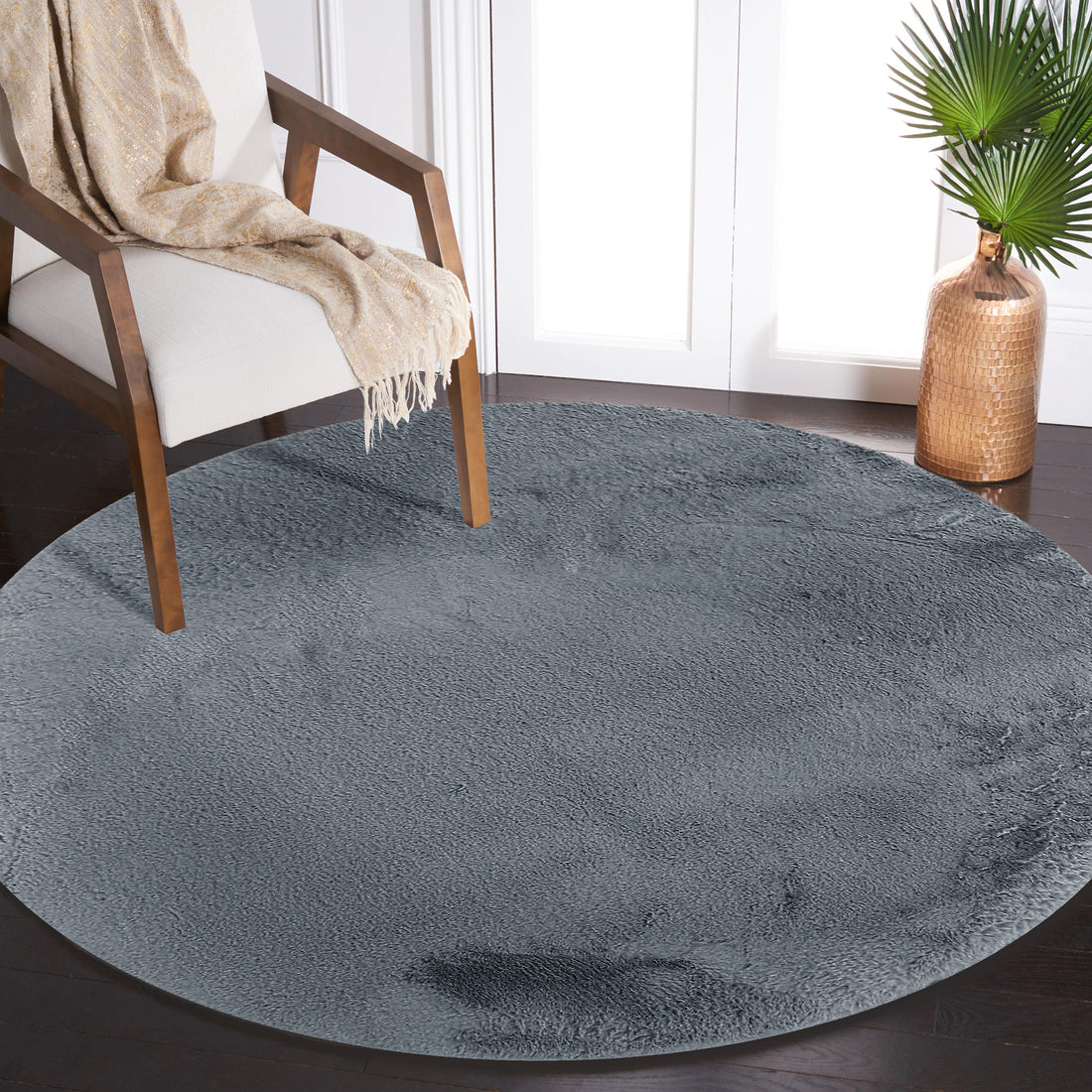 Azaliya Dark Grey Round Faux Rabbit Fur Rug 160cm on dark hardwood, with a mid-century armchair, beige throw, and hammered-metal vase with palm leaves in a sunlit room.