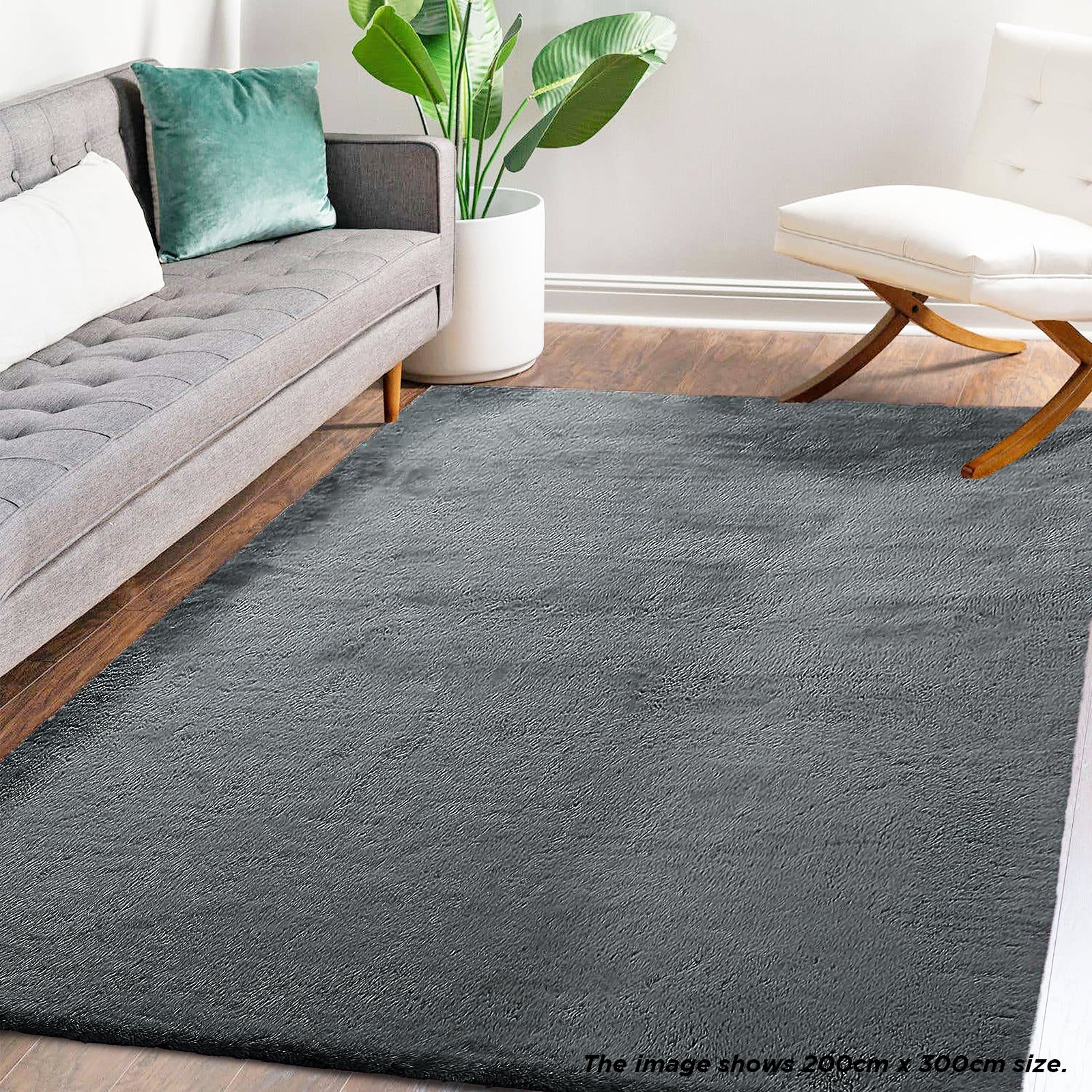 Azaliya Dark Grey Faux Rabbit Fur Rug 200x300cm lies in a modern living room, bordered by a tufted light-gray sofa and a white armless chair, enhancing the cozy, minimalist aesthetic.